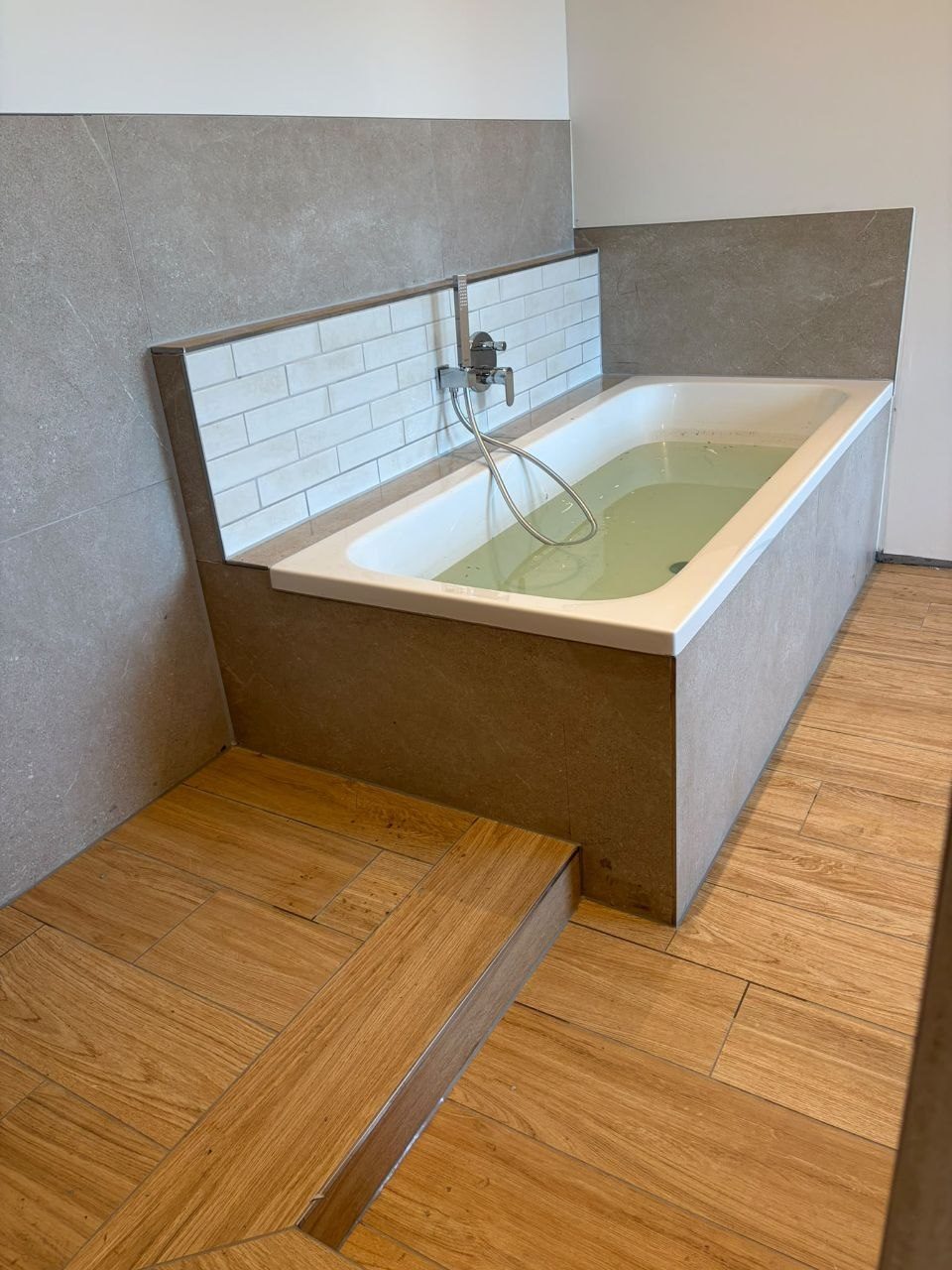 Eingefliest verkleidete Badewanne vor weißen Riemchen im Halbverband, davor ein Podest aus Holzoptik-Fliesen